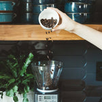 Hand pouring Synastry coffee beans into a grinder with kitchen background and blue ceramic bowls.