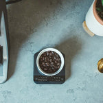 Hypnos decaf coffee beans in a white cup on a digital scale on a gray countertop with a gold spoon and a potted plant nearby