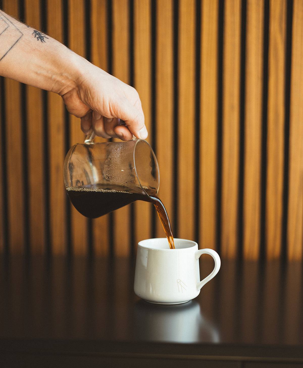 Pouring Synastry coffee blend from glass carafe into white mug on dark table with wooden slat background