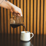 Pouring Synastry coffee blend from glass carafe into white mug on dark table with wooden slat background