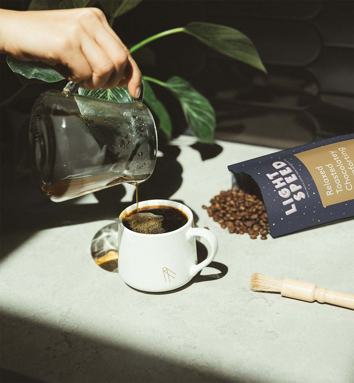Pouring a cup of Hypnos decaf coffee with a bag of Light Speed roasted chocolate coffee beans and coffee grounds brush on a light countertop