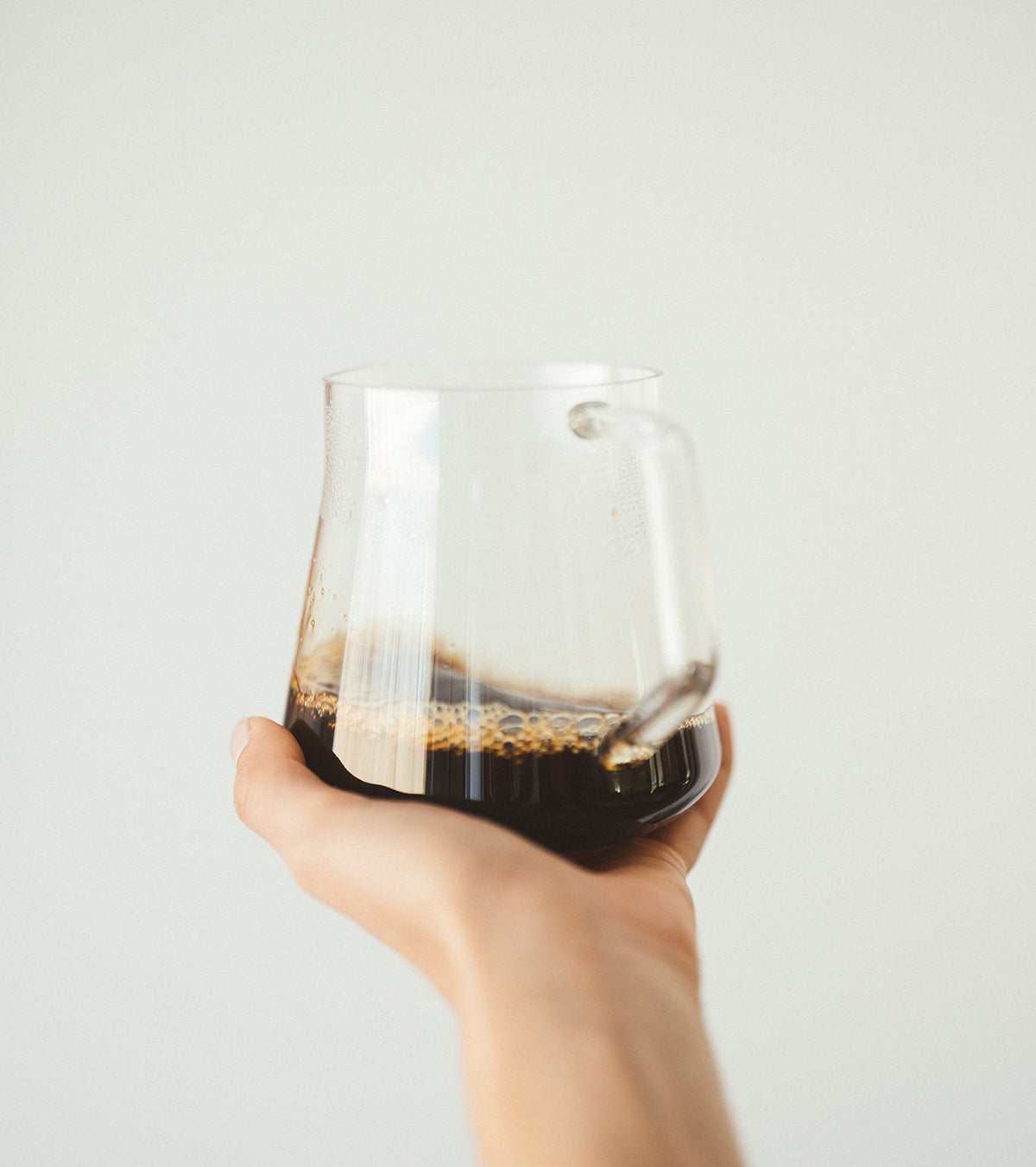 Hand holding a clear glass cup filled with dark Shadow Work coffee against a plain light background