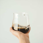 Hand holding a clear glass cup filled with dark Shadow Work coffee against a plain light background