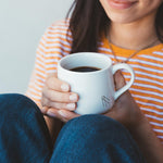 Woman holding a white mug of Hypnos decaf coffee with a striped shirt and jeans