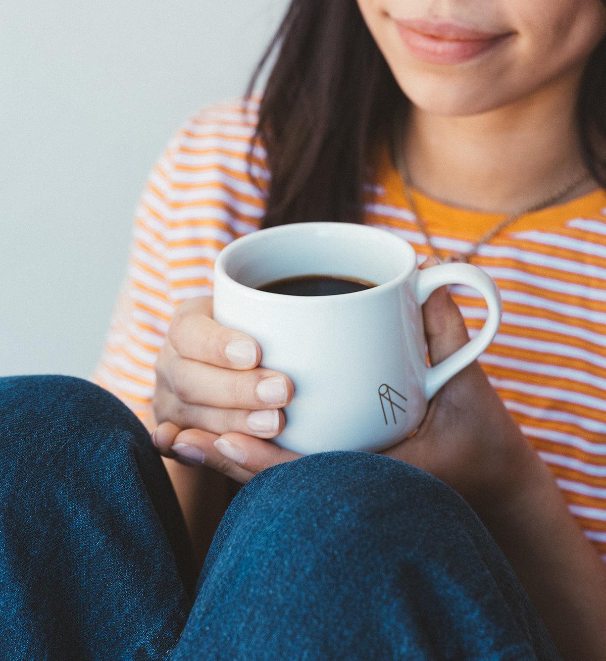 Woman holding a white mug of Hypnos decaf coffee with a striped shirt and jeans